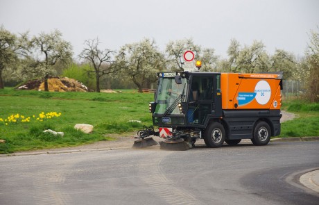 Eine Kehrmaschine mit orange-schwarzer Lackierung reinigt eine gepflasterte Straße in der Nähe einer Wiese mit blühenden Bäumen und gelben Blumen. Im Hintergrund sind Haufen von Schmutz und Steinen zu sehen.