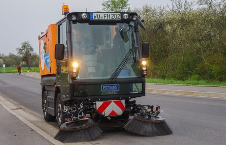 Ein Kehrfahrzeug mit großen rotierenden Besen reinigt den Rand einer gepflasterten Straße. Im Hintergrund sind Bäume und Grünflächen zu sehen, und in der Ferne fährt eine Person auf einem Fahrrad.