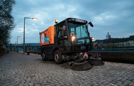 Ein Straßenreinigungsfahrzeug mit großen rotierenden Bürsten kehrt in der Abenddämmerung einen Kopfsteinpflasterweg entlang eines Flusses, wobei der Fahrer im Inneren des Fahrzeugs sitzt und eine Stadt im Hintergrund zu sehen ist.