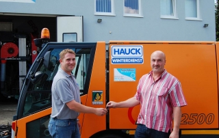 Zwei Männer stehen vor einem orangefarbenen Servicefahrzeug mit der Aufschrift Hauck Winterdienst. Ein Mann übergibt dem anderen die Schlüssel. Beide lächeln und stehen vor einem Gebäude mit Fenstern.