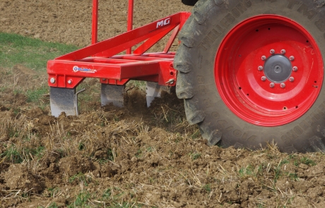 Nahaufnahme eines roten Traktors mit großen Rädern und einem Pflugaufsatz aus Metall, der den Boden auf einem Feld bearbeitet und Erde und Gras umdreht, um die Bepflanzung vorzubereiten.