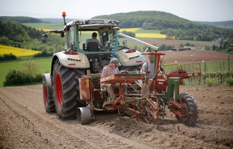 Ein Traktor zieht eine Pflanzmaschine, die von zwei Personen auf einem bestellten Feld bedient wird, mit grünen Hügeln, Bäumen und Ackerland im Hintergrund.
