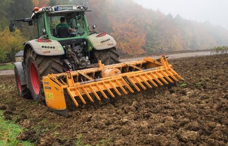 Ein grüner Traktor zieht eine große gelbe Fräse durch ein schlammiges Feld und bearbeitet den Boden. Im Hintergrund erscheinen Herbstbäume mit buntem Laub und leichtem Nebel.