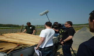Ein Filmteam mit Kameras und Galgenmikrofonen nimmt einen Mann auf, der an einem mit Holzbrettern beladenen Anhänger an einer Landstraße steht, mit Feldern und blauem Himmel im Hintergrund.