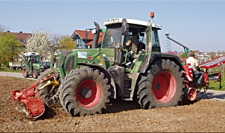 Ein grüner Traktor mit großen Reifen pflügt an einem sonnigen Tag ein Feld. Im Hintergrund sind einige Menschen und ein weiterer Traktor in der Nähe einiger Häuser und Bäume zu sehen.
