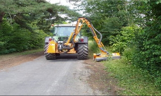 Ein Traktor mit angebautem Mäharm stutzt die Vegetation am Straßenrand und pflegt das Grün an der von Bäumen und Büschen gesäumten Landstraße.