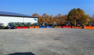 Eine Reihe von Traktoren mit farbenfrohen Schneepflügen (rot, blau, orange, gelb) sind draußen auf einem Schotterplatz in der Nähe eines großen Gebäudes und von Bäumen mit Herbstlaub geparkt. Der Himmel ist klar und blau.