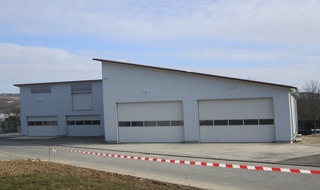 Ein großes, modernes Industriegebäude mit drei breiten Garagentoren und einem schrägen Dach steht neben einer gepflasterten Straße, die von einer rot-weiß gestreiften Absperrung begrenzt wird, unter einem teilweise bewölkten Himmel.