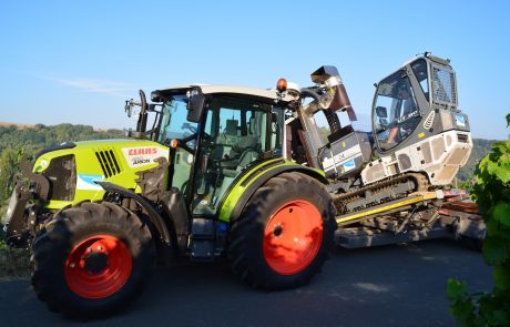 Ein grün-schwarzer CLAAS Arion Traktor mit roten Rädern transportiert eine große Raupenlandmaschine auf einem Tieflader im Freien bei Tageslicht. Im Hintergrund sind Bäume und Hügel zu sehen.