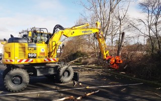 Ein gelber Komatsu-Bagger mit einem Baumschneidegerät schneidet Äste von Bäumen in der Nähe eines Parkplatzes ab, wobei die abgeschnittenen Äste auf dem Boden verstreut liegen. Der Himmel ist teilweise bewölkt.