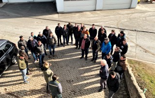 Eine Gruppe von etwa 25 Personen steht in einem Halbkreis auf einer gepflasterten Fläche im Freien und hört zwei Personen zu, die zu ihnen sprechen. Im Hintergrund sind mehrere Garagentore und Fahrzeuge zu sehen.