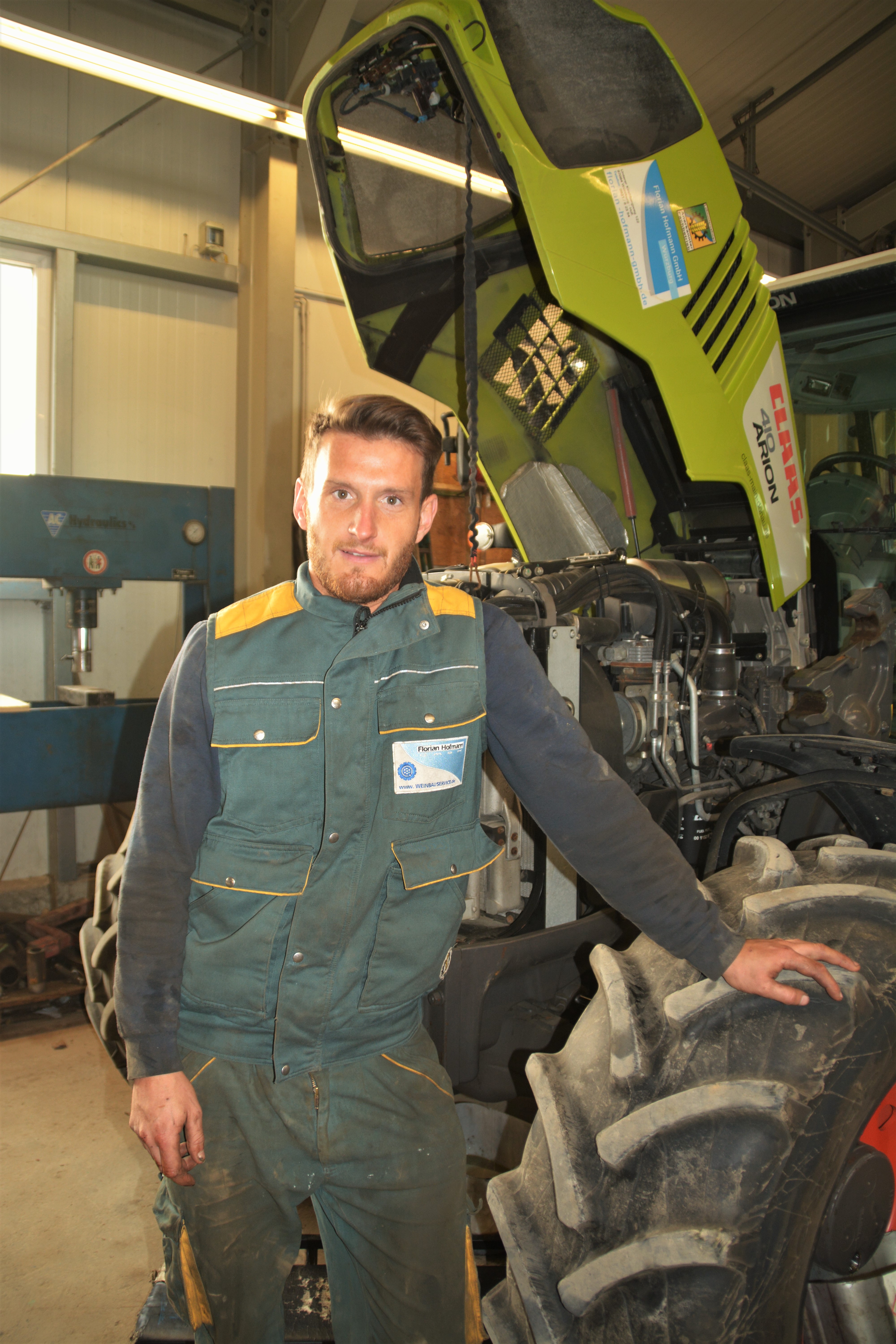 Ein Mann in einem Arbeitsoverall steht neben einem großen Traktor in einer Werkstatt. Die Motorhaube des Traktors ist geöffnet, und der Mann stützt sich mit einer Hand auf einen Vorderreifen und schaut in die Kamera. Ein Mann in einem Arbeitsoverall steht neben einem großen Traktor in einer Werkstatt. Die Motorhaube des Traktors ist geöffnet, und der Mann stützt sich mit einer Hand auf einen Vorderreifen und schaut in die Kamera.