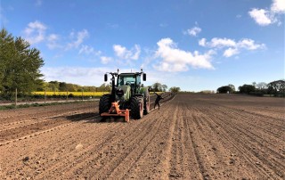 Ein Traktor fährt über ein großes, frisch gepflügtes Feld unter einem blauen Himmel mit vereinzelten Wolken. Bäume und Grünflächen begrenzen das Feld im Hintergrund.