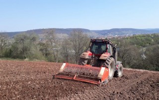 Ein roter Traktor pflügt ein Feld an einem Hang, im Hintergrund sind Bäume und eine Stadt zu sehen, unter einem klaren blauen Himmel.