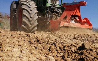Nahaufnahme eines großen grünen Traktors mit einer roten Bodenfräse, der unter blauem Himmel trockenen, felsigen Boden pflügt. Hinter dem Traktor steigt Staub auf, während er sich vorwärts bewegt.