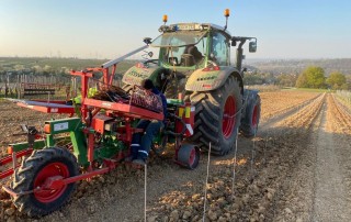 Ein Traktor mit angehängten Maschinen arbeitet auf einem gepflügten Feld unter freiem Himmel. Eine Person sitzt auf der Maschine, und es sind Reihen von Erde mit Pflöcken zu sehen, die auf Pflanzungen oder laufende landwirtschaftliche Arbeiten hindeuten.