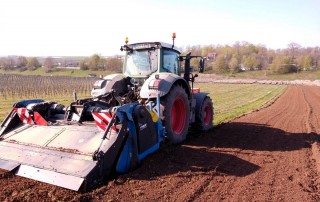 Ein Traktor mit großen Hinterrädern und angehängten landwirtschaftlichen Geräten bearbeitet den Boden auf einem Feld mit Reihen von Bäumen im Hintergrund unter einem klaren Himmel.