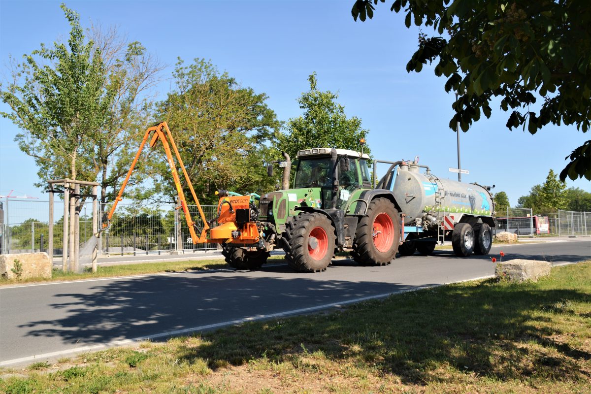 Ein Traktor mit einem großen Tankanhänger und orangefarbenem Anbaugerät bewässert an einem sonnigen Tag junge Bäume am Straßenrand, während im Hintergrund Grünflächen und Zäune zu sehen sind. Ein Traktor mit einem großen Tankanhänger und orangefarbenem Anbaugerät bewässert an einem sonnigen Tag junge Bäume am Straßenrand, während im Hintergrund Grünflächen und Zäune zu sehen sind.