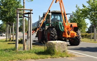 Ein Traktor mit einem großen Tank und einem angebrachten orangefarbenen Arm bewässert an einem sonnigen Tag junge Bäume, die entlang einer gepflasterten Straße gepflanzt wurden. Die Szene zeigt grünes Gras, kleine Bäume und einen Steinblock in der Nähe des Bordsteins.