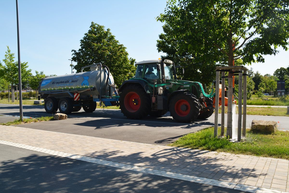 Ein grüner Traktor, der einen silbernen Tankanhänger zieht, fährt an einem sonnigen Tag eine gepflasterte Straße entlang, die von jungen Bäumen gesäumt ist. In der Nähe sind Gras und ein paar Felsen zu sehen, und der Himmel ist klar. Ein grüner Traktor, der einen silbernen Tankanhänger zieht, fährt an einem sonnigen Tag eine gepflasterte Straße entlang, die von jungen Bäumen gesäumt ist. In der Nähe sind Gras und ein paar Felsen zu sehen, und der Himmel ist klar.