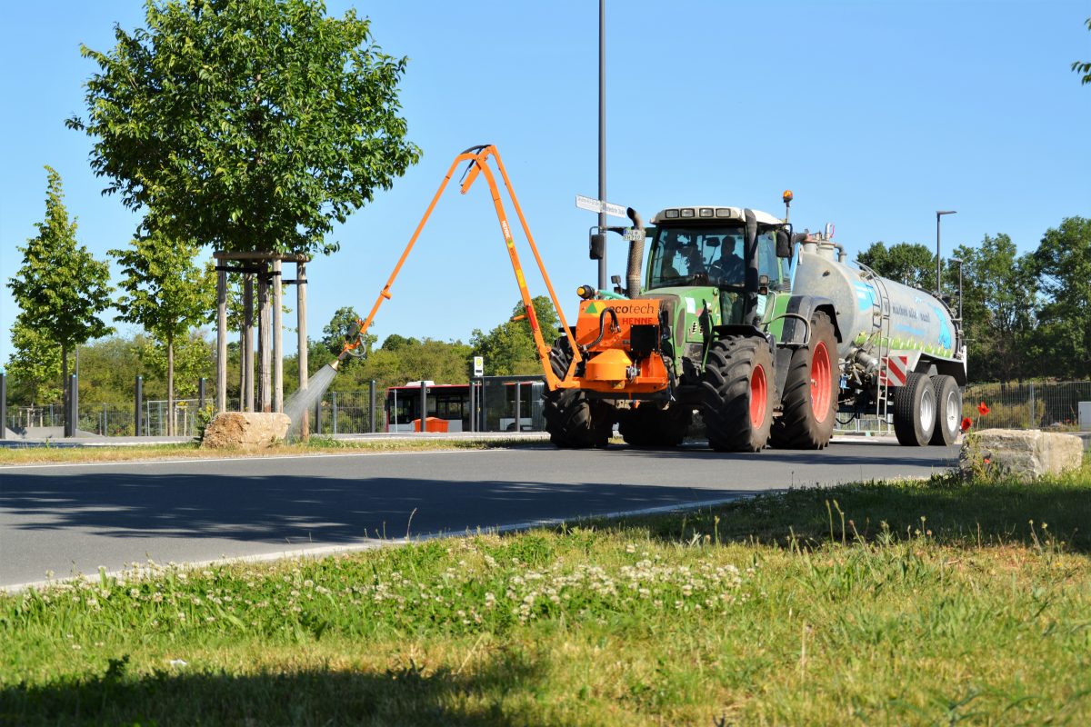 Ein Traktor mit einem großen Tank und einem angebrachten Arm besprüht einen neu gepflanzten Baum am Straßenrand in einer sonnigen, grünen Gegend. Im Hintergrund sind Gras, Bäume und ein klarer blauer Himmel zu sehen. Ein Traktor mit einem großen Tank und einem angebrachten Arm besprüht einen neu gepflanzten Baum am Straßenrand in einer sonnigen, grünen Gegend. Im Hintergrund sind Gras, Bäume und ein klarer blauer Himmel zu sehen.