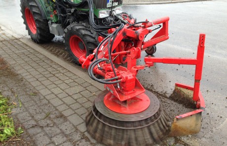 Ein grüner Traktor mit einem großen roten rotierenden Bürstenaufsatz fegt Schmutz und Geröll vom Rand eines Kopfsteinpflasters auf die Straße.