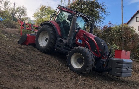 Ein roter Traktor mit großen Reifen fährt auf einem steilen, grasbewachsenen Hügel. Im Hintergrund sind Bäume und ein Haus zu sehen, und der Traktor scheint das abschüssige Gelände zu bearbeiten.