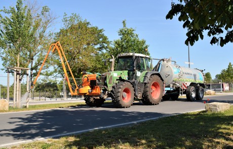 Ein Traktor mit einem großen Wassertank und einem angebrachten orangefarbenen Arm bewässert an einem sonnigen Tag Bäume am Straßenrand. Gras und Bäume säumen die Straße, der Himmel ist klar und blau.