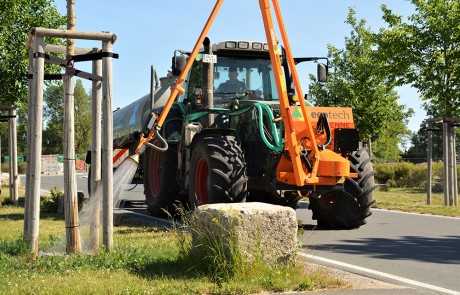 Ein Traktor mit einem großen Wassertank bewässert mit einem ausziehbaren Schlauch und einer Düse die neu gepflanzten Bäume an einem Straßenrand. Die Bäume werden von Holzpfählen gestützt, und die Szene ist sonnig mit klarem Himmel.