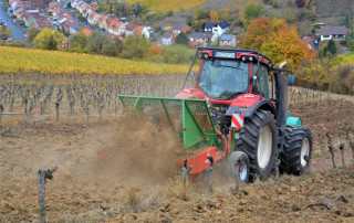 Ein roter Traktor pflügt einen Weinberg am Hang und wirbelt die Erde zwischen den Reihen der beschnittenen Rebstöcke auf. Im Hintergrund sind ein Dorf und herbstlich gefärbte Bäume zu sehen.