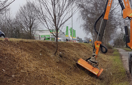 Ein Arbeiter bedient einen an einem Fahrzeug befestigten orangefarbenen Rasenmäher und mäht Gras an einer schrägen Böschung neben einer Straße. Im Hintergrund sind blattlose Bäume, ein blaues geparktes Auto und eine Tankstelle zu sehen.