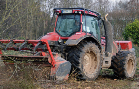 Ein roter Traktor mit großen, schlammigen Reifen beseitigt mit einem Frontanbaugerät Gestrüpp und kleine Bäume in einer ländlichen Umgebung. Im Hintergrund sind Bäume und dichte Vegetation zu sehen.