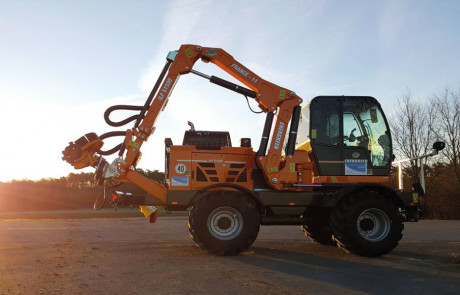 Ein orangefarbenes Baufahrzeug mit einem ausgefahrenen Hydraulikarm steht bei Sonnenuntergang auf einer gepflasterten Fläche, im Hintergrund sind Bäume und offene Felder zu sehen.
