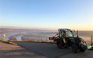 Ein Traktor ist auf einem gepflasterten Weg geparkt und überblickt ein nebliges Tal mit Feldern, Bäumen und Häusern, die in der Ferne unter einem klaren blauen Himmel bei Sonnenaufgang oder Sonnenuntergang zu sehen sind.