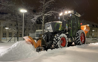Ein Schneepflugtraktor mit hellen Lichtern räumt nachts frischen Schnee auf einer Straße, umgeben von Gebäuden und schneebedeckten Bäumen. Der Schnee fällt sanft und schafft eine heitere Winterszene.
