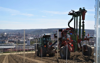 Ein Traktor mit angehängten landwirtschaftlichen Geräten arbeitet auf einem bearbeiteten Feld in Hanglage mit Blick auf eine Stadtlandschaft mit Gebäuden und entfernten Hügeln unter einem klaren blauen Himmel.