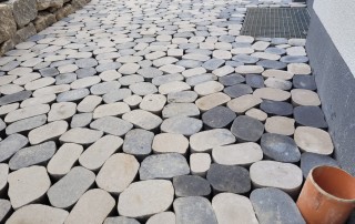 Ein steinerner Weg aus unregelmäßig geformten, hellen und dunklen Pflastersteinen führt an einer Steinmauer und einem Gebäude entlang. Auf dem Boden befindet sich ein Metallgitter und in der rechten unteren Ecke ein braunes Rohr.