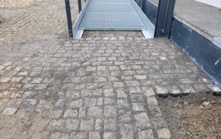Eine behindertengerechte Metallrampe führt hinunter zu einem unebenen Kopfsteinpflaster, das an den Rändern des Weges einige schmutzige und unfertige Stellen aufweist.