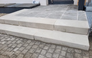 Betontreppen mit drei breiten, flachen Setzstufen führen zu einem gefliesten Podest vor einem Gebäudeeingang. Der Bereich unterhalb der Stufen ist mit Pflastersteinen gepflastert. Auf der linken Seite ist ein kleines Schild zu sehen.