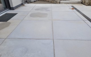 Große, hellgraue Betonplatten mit einigen feuchten Stellen, die von einer Gebäudemauer und Entwässerungsgittern begrenzt werden, mit Steinblöcken und unvollendeter Landschaftsgestaltung im Hintergrund.