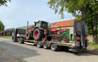 Ein großer Pritschenwagen ist auf einer Kopfsteinpflasterstraße geparkt und transportiert einen rot-grünen Traktor mit angehängten landwirtschaftlichen Geräten. Im Hintergrund sind Scheunen und Bäume zu sehen.