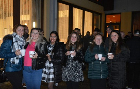 Eine Gruppe von sechs Frauen steht nachts draußen, lächelt und hält weiße Tassen in der Hand. Sie sind warm mit Mänteln und Schals gekleidet. Im Hintergrund sind ein beleuchtetes Gebäude und einige andere Menschen zu sehen.