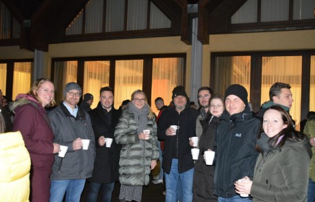 Eine Gruppe warm gekleideter Menschen in Mänteln und Mützen steht nachts vor einem Gebäude, hält Tassen in der Hand und lächelt in die Kamera. Die Fenster des Gebäudes sind von innen beleuchtet und bilden einen gemütlichen Hintergrund.