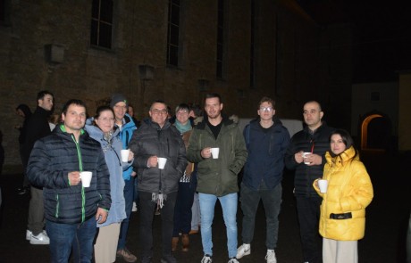 Eine Gruppe von Menschen steht nachts im Freien, hält weiße Becher in der Hand und trägt warme Jacken. Sie stehen vor einem Steingebäude mit hohen Fenstern, und die Atmosphäre wirkt entspannt und freundlich.