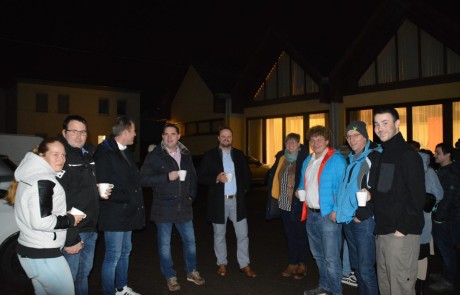 Eine Gruppe von Menschen steht nachts draußen, warm gekleidet in Jacken und mit Tassen in der Hand, lächelnd und gesellig vor Gebäuden mit warm beleuchteten Fenstern.