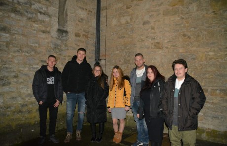 Sieben Personen stehen in einem schwach beleuchteten Bereich mit Steinmauern zusammen und posieren für ein Gruppenfoto. Die meisten tragen dunkle Jacken, eine Person in der Mitte hat eine gelbe Jacke an. Sie schauen alle in die Kamera und lächeln.