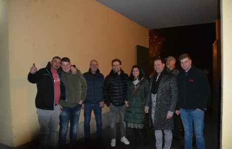 Eine Gruppe von neun winterlich gekleideten Personen steht lächelnd in einem schwach beleuchteten Flur mit gelben Wänden zusammen. Einige haben die Hände in den Taschen, und eine Person auf der linken Seite zeigt den Daumen nach oben.