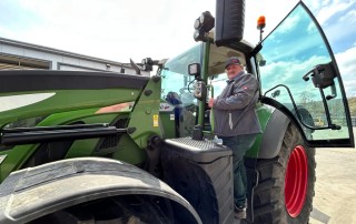 Eine Person in Jacke und Mütze steht auf den Stufen eines großen grünen Traktors, hält die Tür offen und lächelt in die Kamera. Der Traktor ist im Freien in der Nähe eines Gebäudes unter einem teilweise bewölkten Himmel geparkt.