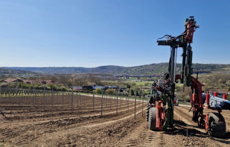 Eine große Landmaschine steht in einem bestellten Feld mit Pfahlreihen, wahrscheinlich für einen Weinberg, unter einem klaren blauen Himmel. Im Hintergrund sind Hügel, Häuser und eine entfernte Brücke zu sehen.