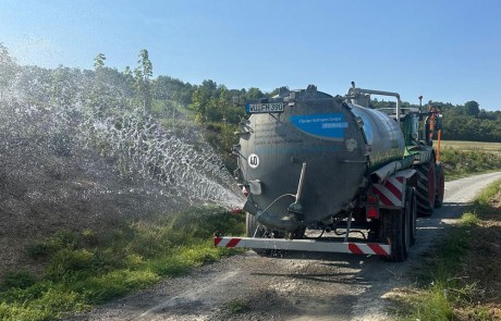Ein Traktor zieht einen Tank, der von hinten eine Flüssigkeit auf ein Feld sprüht. Er fährt auf einem ländlichen Feldweg, der von grünem Gras und Bäumen gesäumt ist, unter einem klaren blauen Himmel.